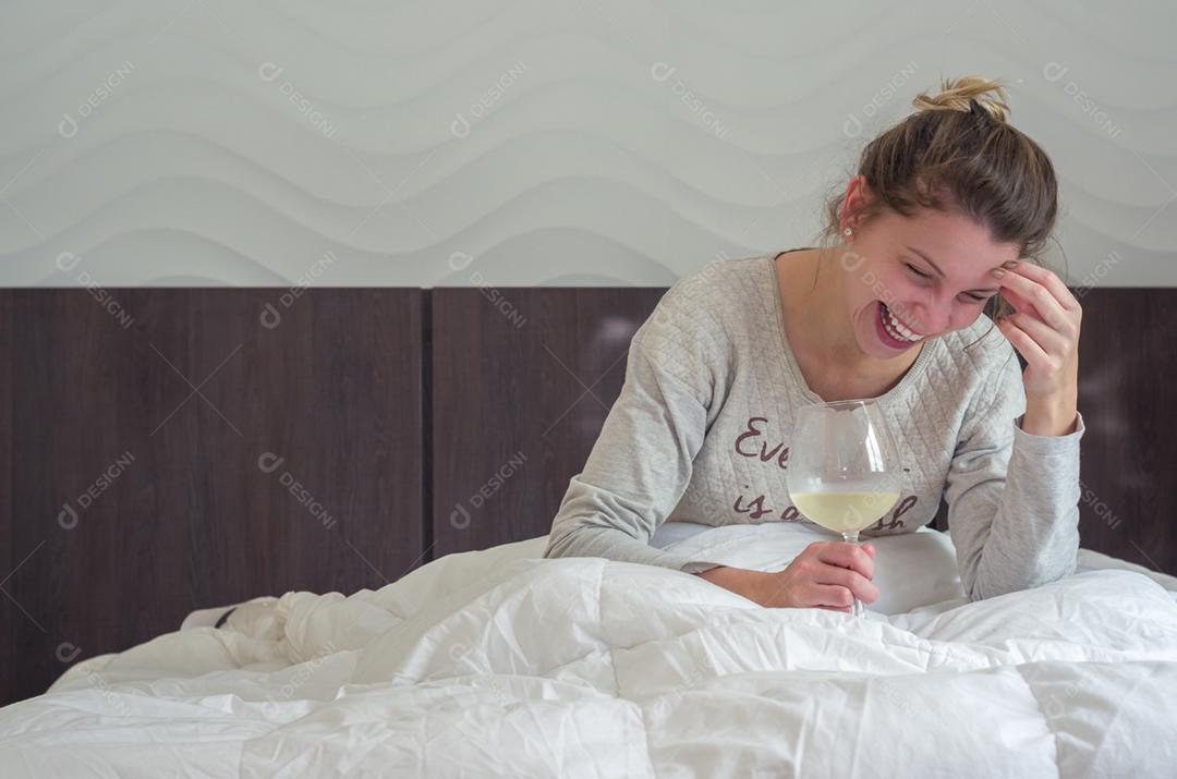 Jovem loira rindo na cama com um copo de vinho na mão, acordando feliz.