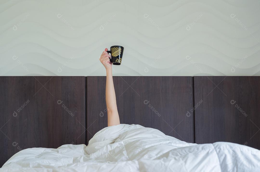 Tomando café amanhã cedo. Mulher na cama, debaixo do edredom, com o braço levantado segurando a xícara de café.