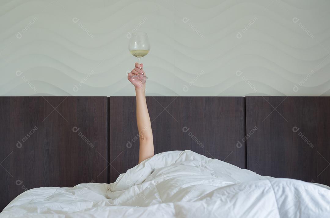 Mulher na cama, debaixo do edredom, com o braço levantado segurando o copo de vinho