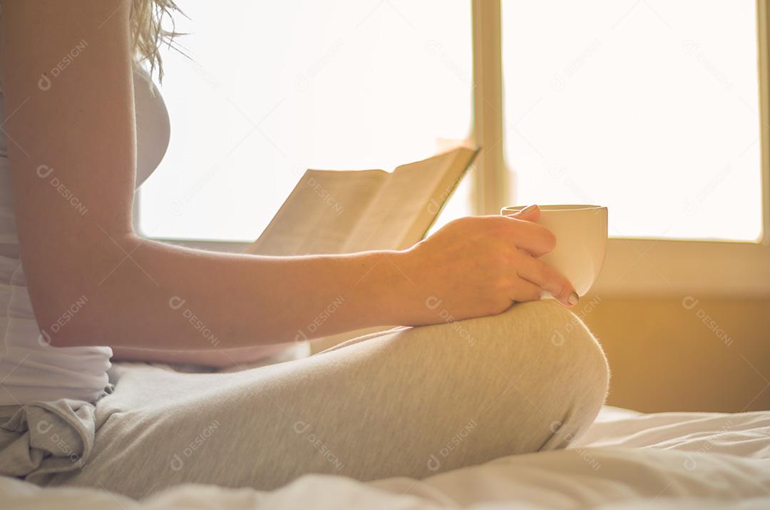 Mulher lendo livro, tomando café na cama com raios solares em th