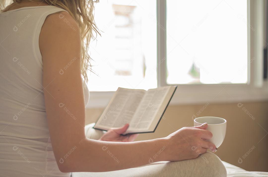 Mulher lendo livro, tomando café na cama com raios solares em th