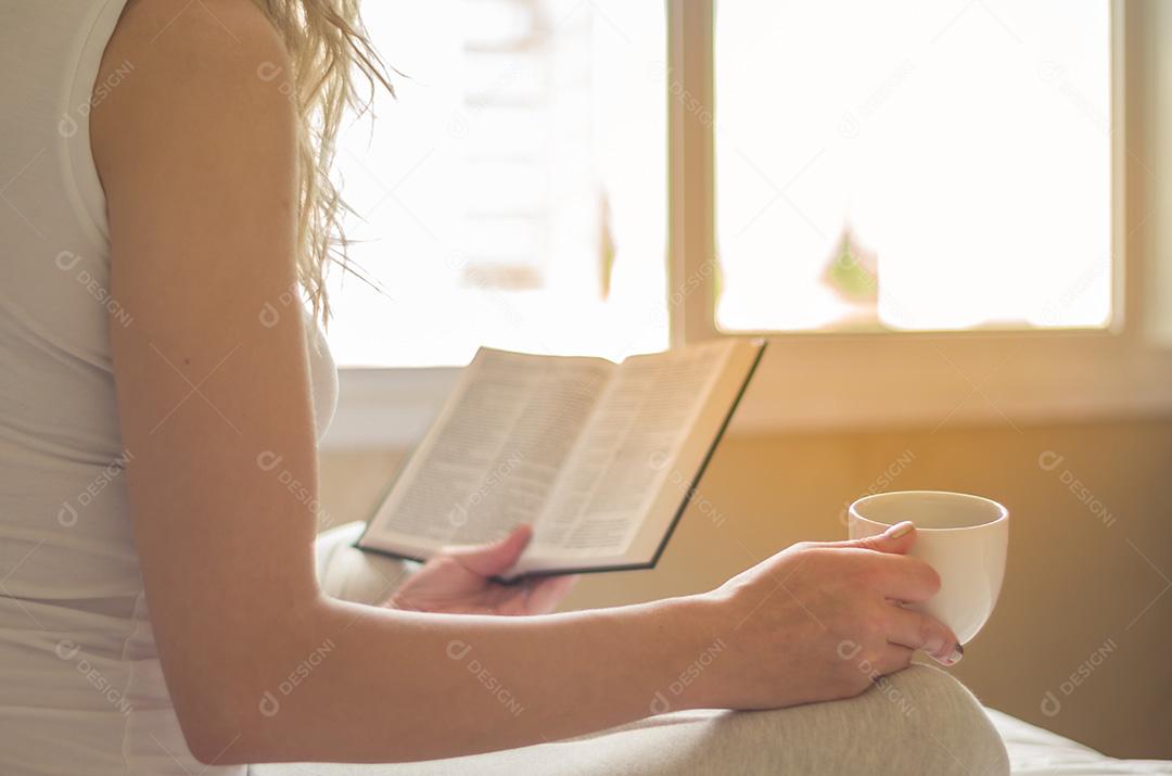 Mulher lendo livro, tomando café na cama com raios solares em th