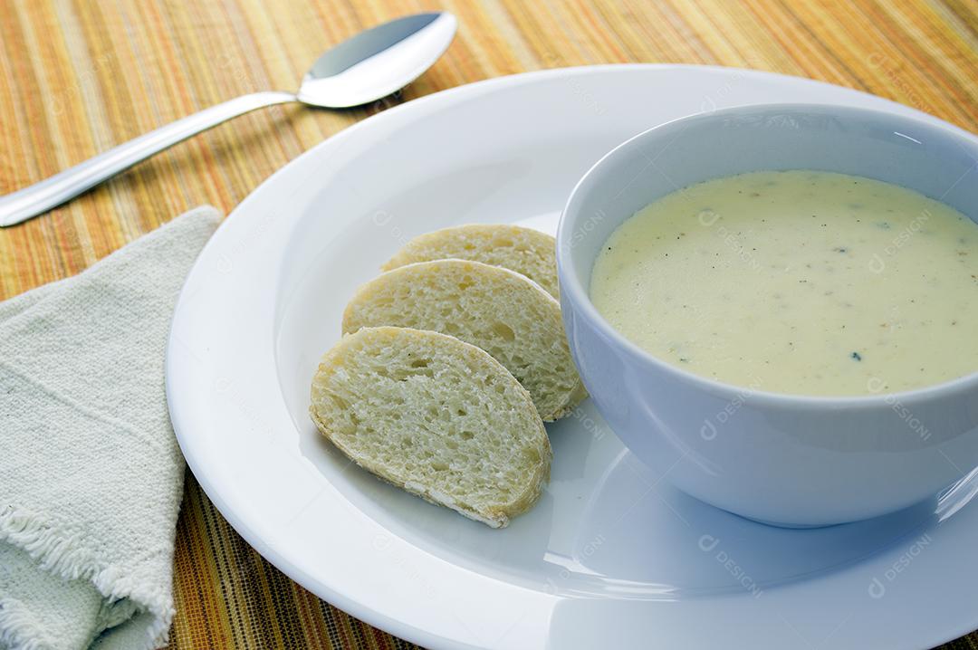 Sopa de queijo em uma tigela branca sobre um prato com pão fatiado.