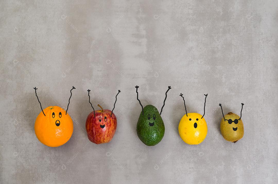 Diversas frutas com rostos felizes e braços para cima em fundo cinza, concreto polido. Laranja, maçã, kiwi, limão.