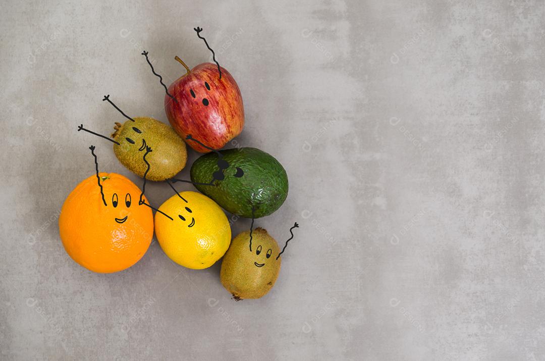 Diversas frutas com rostos felizes e braços para cima em fundo cinza, concreto polido. Laranja, maçã, kiwi, limão.