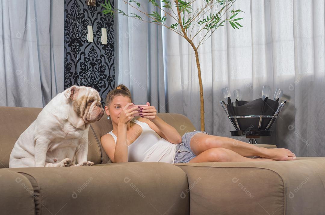 Jovem mulher usando smartphone no sofá com cachorro de estimação (buldogue inglês)