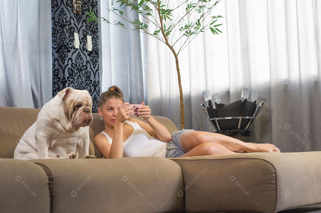 Jovem mulher usando smartphone no sofá com cachorro de estimação (buldogue inglês)