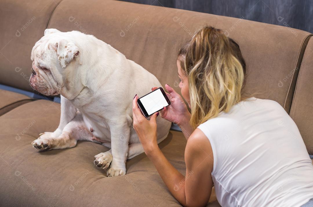 Jovem mulher usando smartphone no sofá com cachorro de estimação (buldogue inglês)