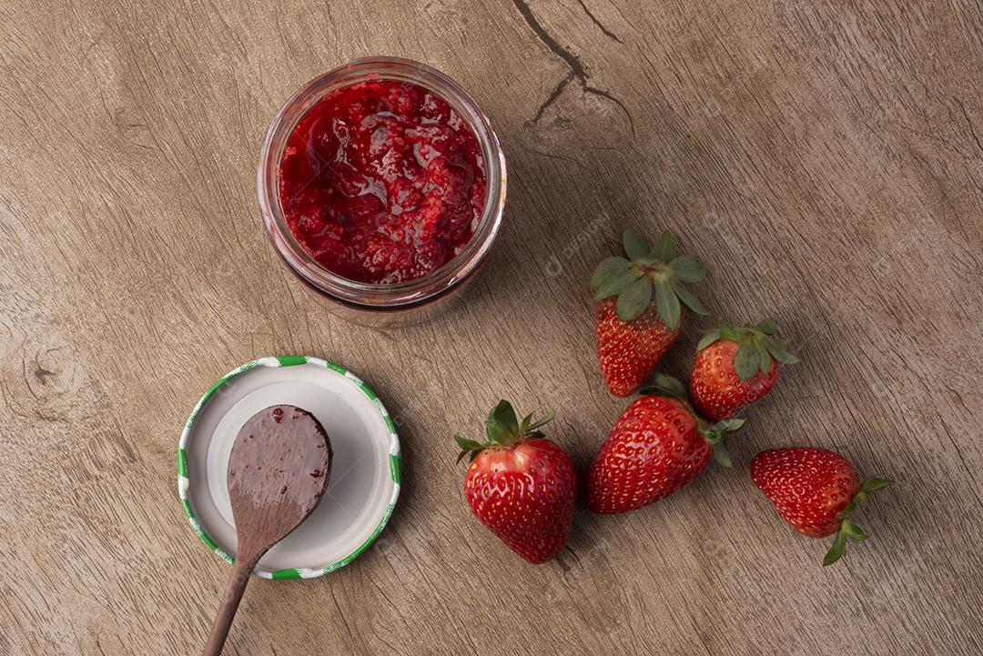 Copo de geleia de morango, colher e morangos sobre mesa de madeira.