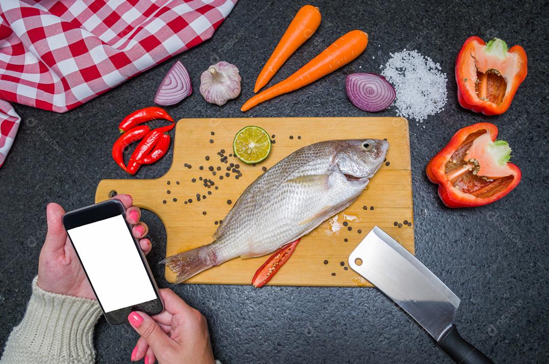 Receita de consultoria jovem para peixe no smartphone, peixe e ingredientes na mesa.