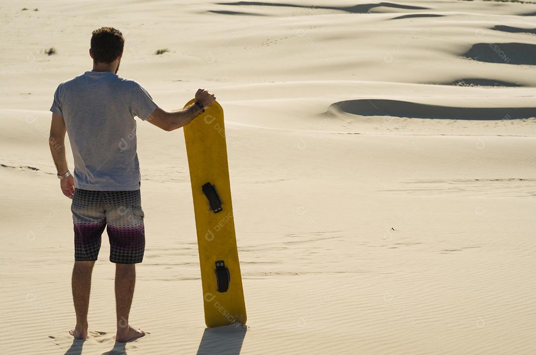 Jovem de costas olhando para as dunas de areia, preparando-se para praticar sandboard.