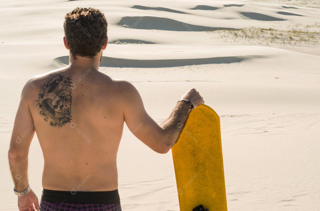 Jovem de costas olhando para as dunas de areia, preparando-se para praticar sandboard.