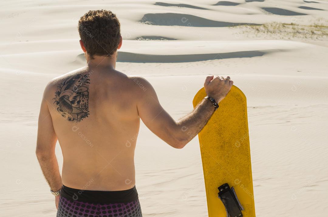 Jovem de costas olhando para as dunas de areia, preparando-se para praticar sandboard.
