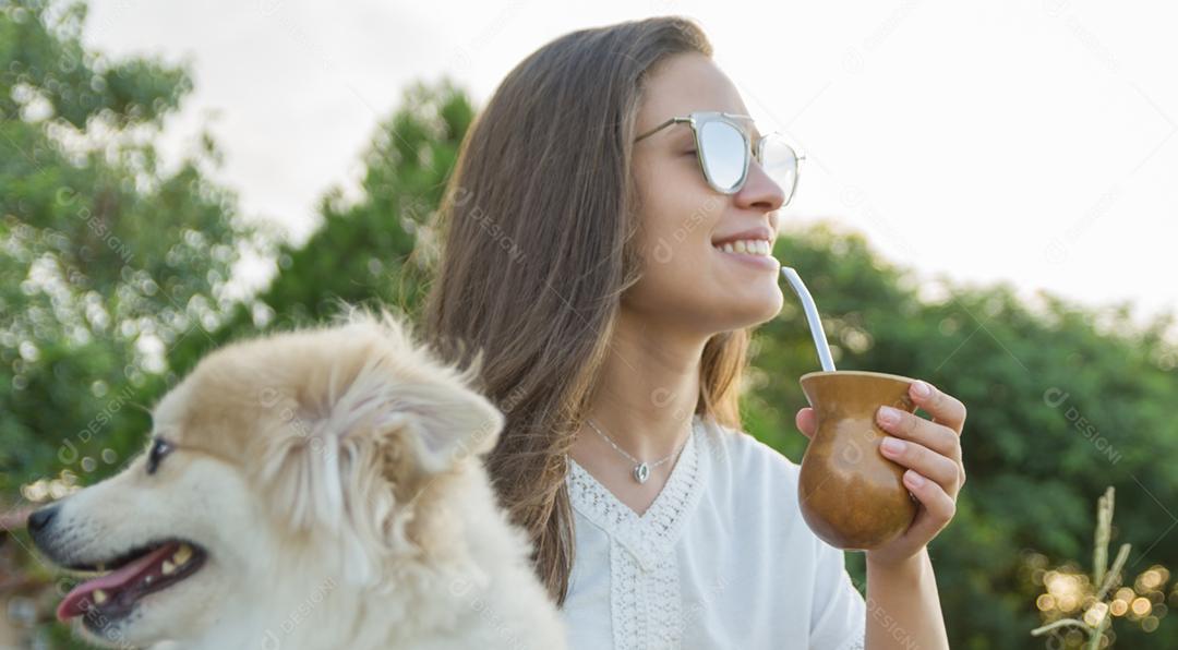 Mulher loira tomando chimarrao tradicional gaúcho com cachorro no parque.