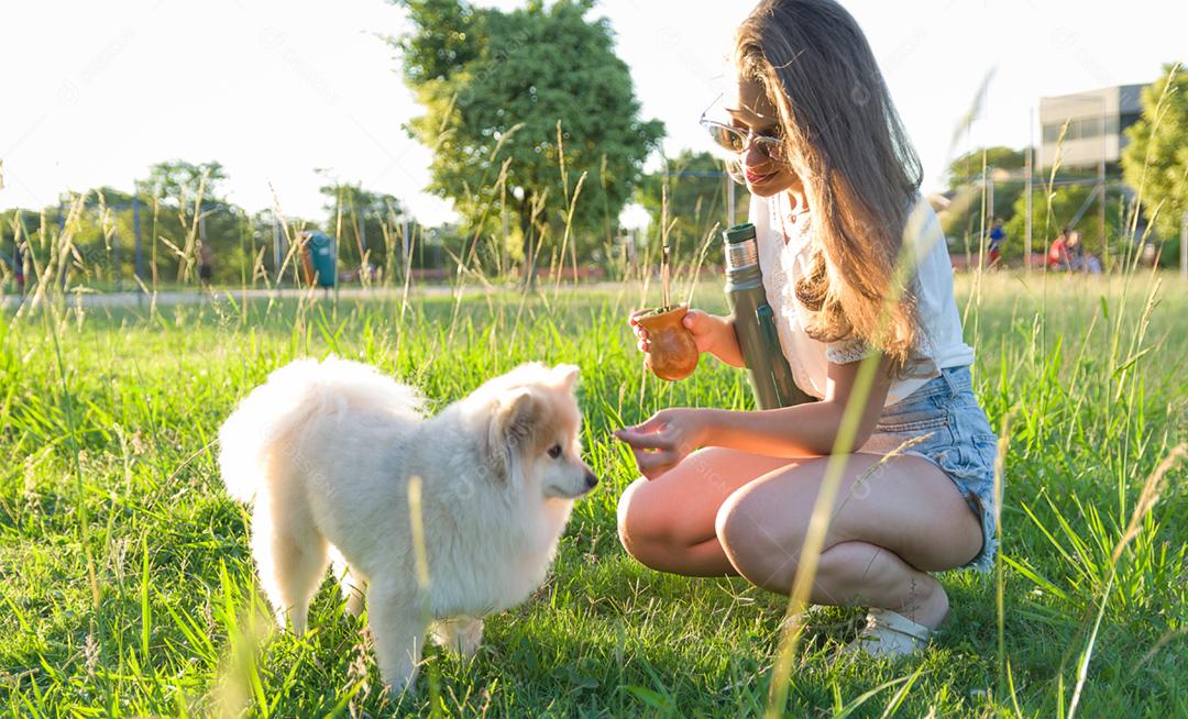 Mulher loira tomando chimarrao tradicional gaúcho com cachorro no parque.