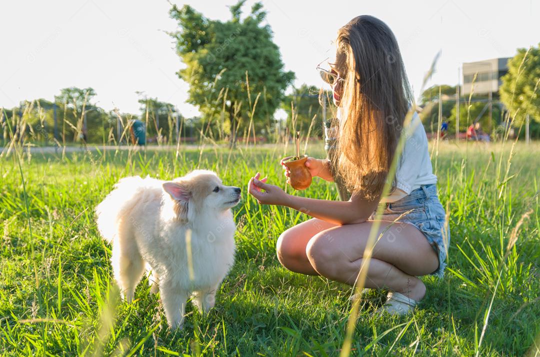 Mulher loira tomando chimarrao tradicional gaúcho com cachorro no parque.