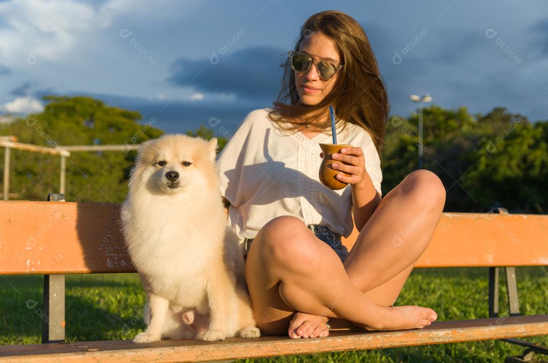 Mulher loira tomando chimarrao tradicional gaúcho com cachorro no parque.