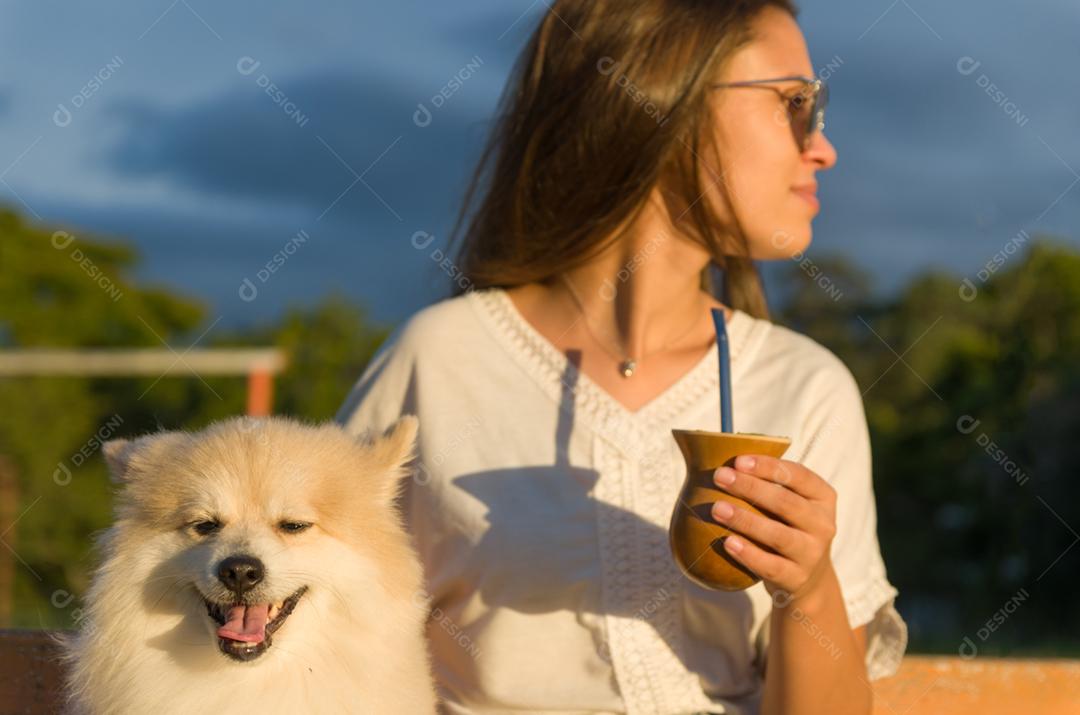Mulher loira tomando chimarrao tradicional gaúcho com cachorro no parque.