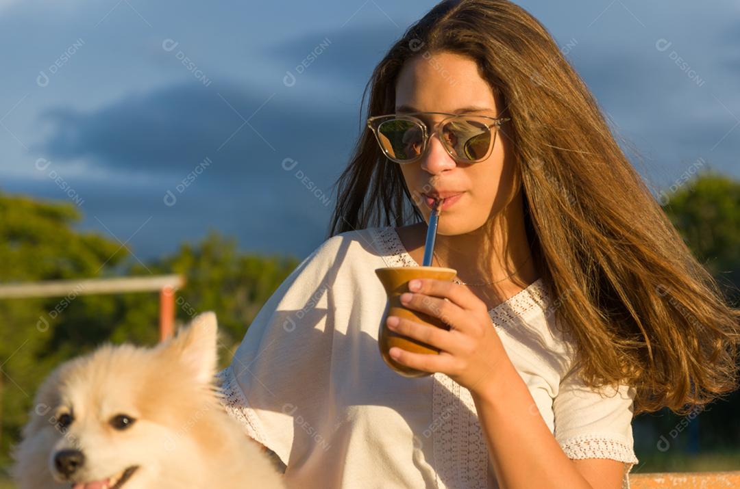 Mulher loira tomando chimarrao tradicional gaúcho com cachorro no parque.