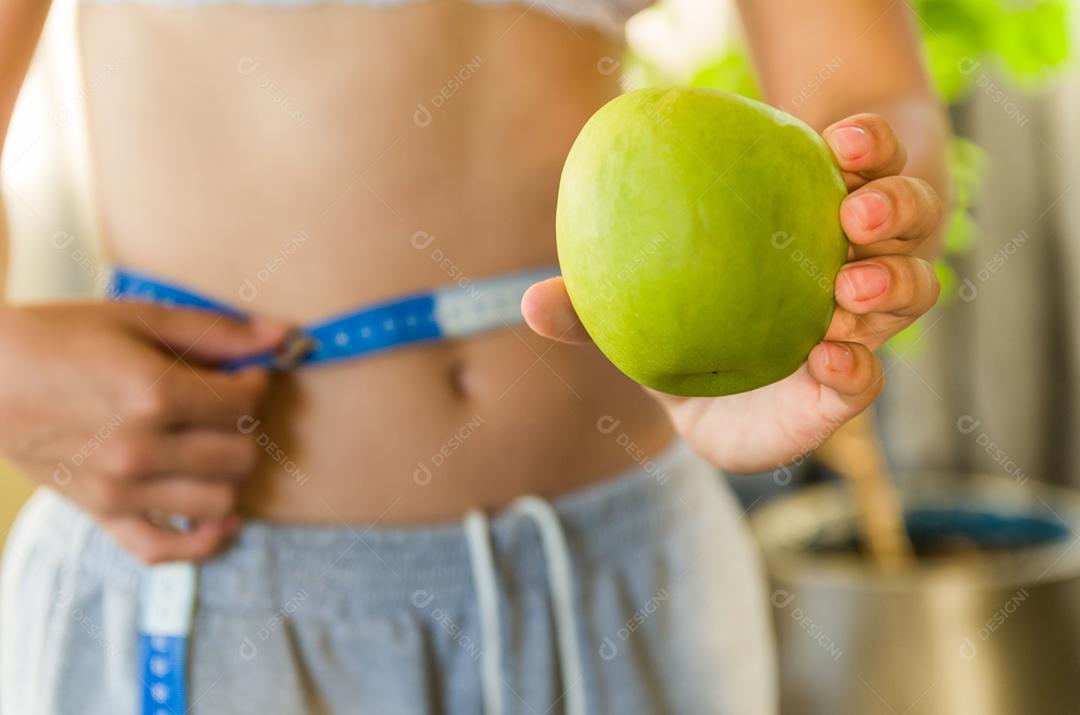 Mulher jovem magra medindo sua cintura fina com uma fita métrica e mostrando uma maçã
