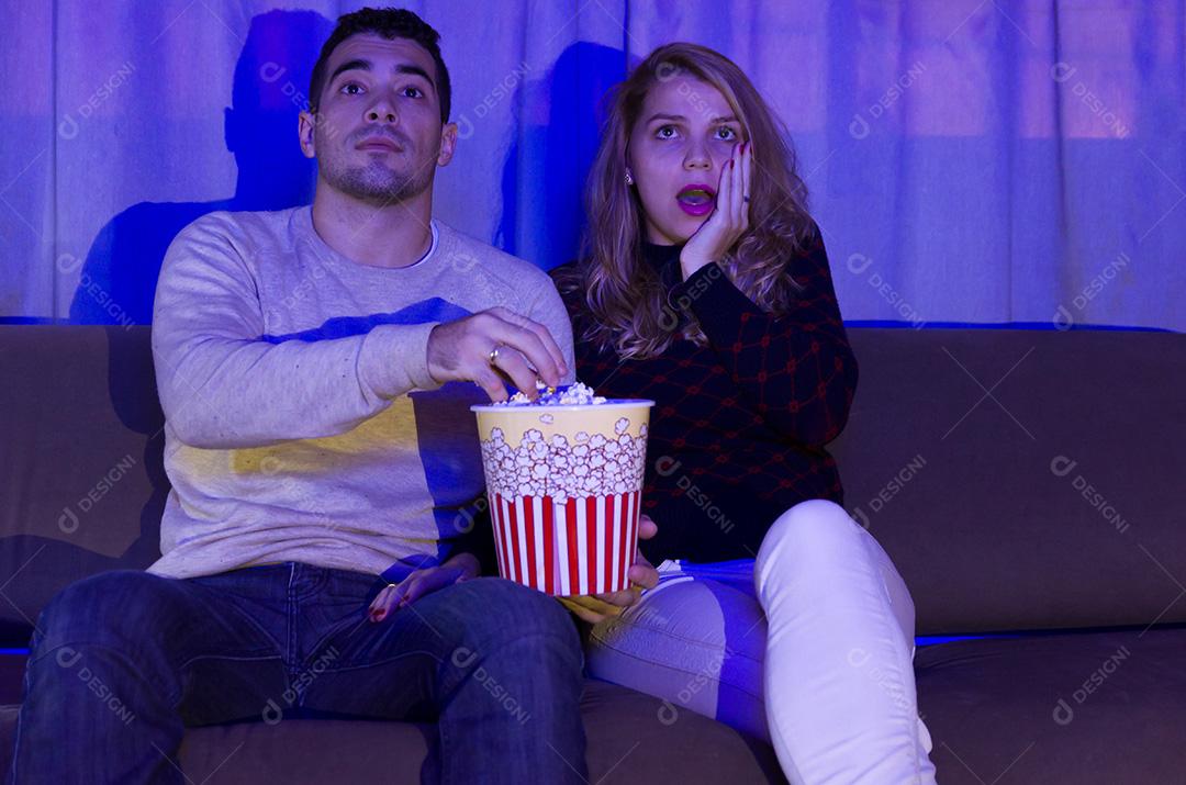 Jovem casal assistindo filme de suspense no quarto escuro com pipoca o