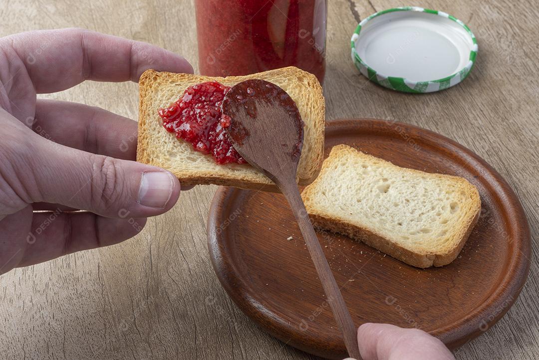 Mão segurando torrada e colher com geléia de morango