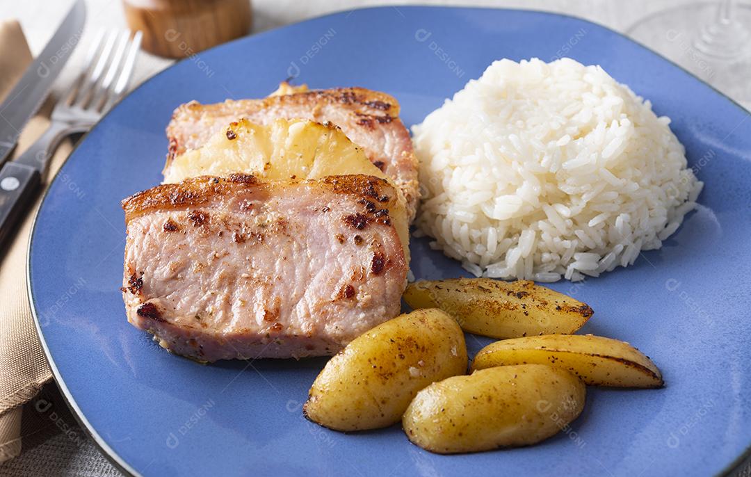 Estacas de porco com abacaxi, arroz e batatas sobre prato azul.