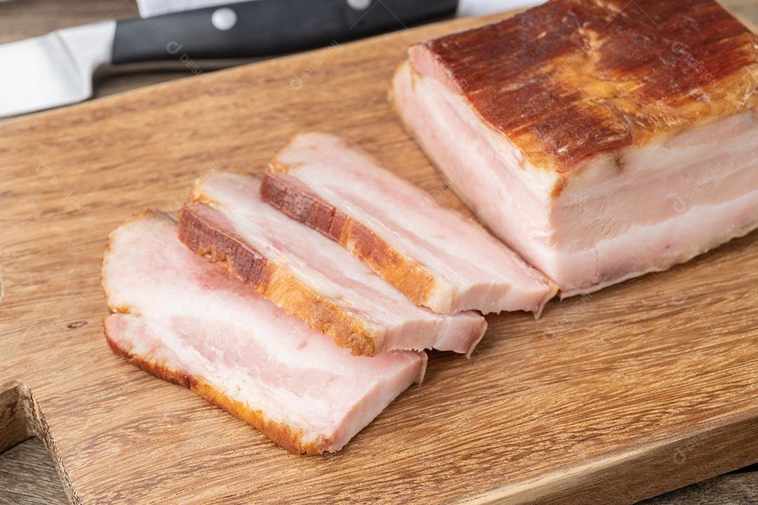 Bacon defumado com fatias e faca sobre placa de madeira.