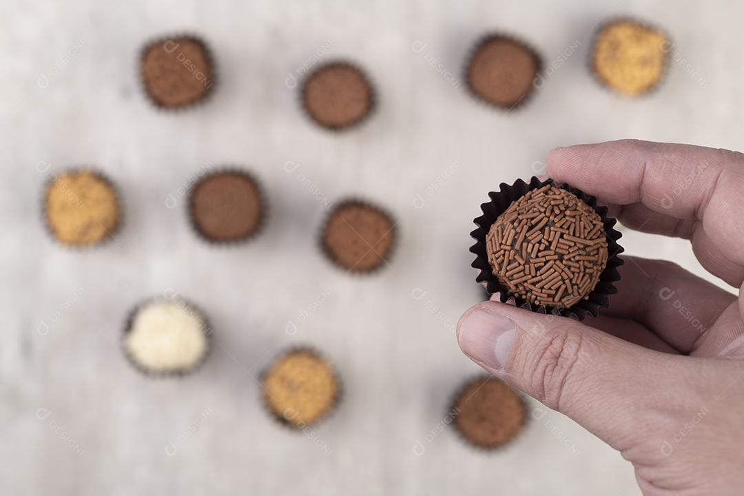 Mão segurando brigadeiros típicos brasileiros sobre brigadeiros desfocados no fundo.