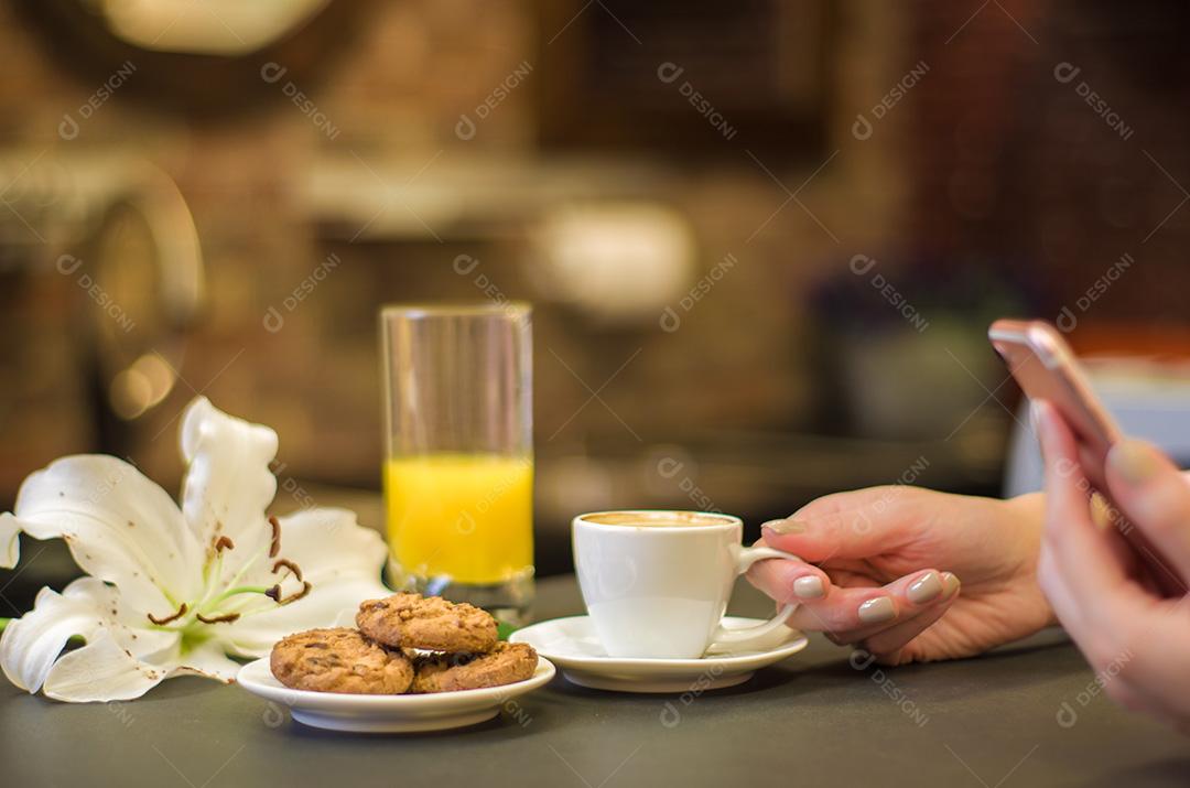 Mesa posta, café , biscoitos e suco com fundo escuro mulher segurando xicara olhando celular
