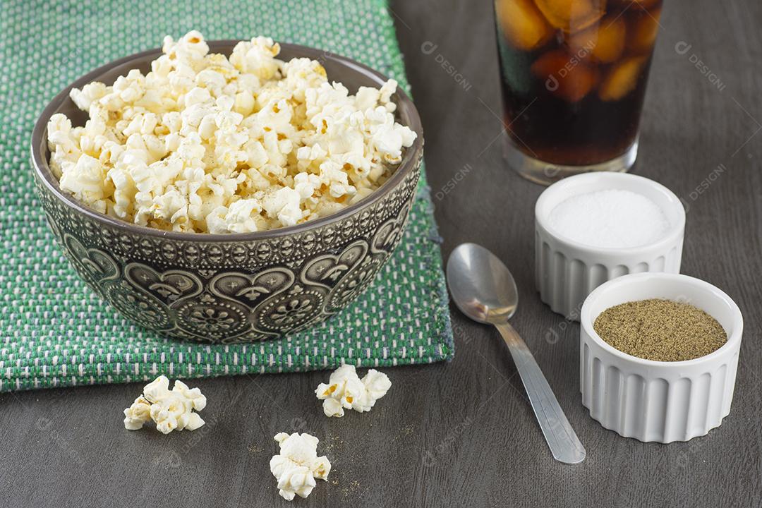 Pipocas em uma tigela decorada com sal uma pimenta e um copo de refrigerante sobre uma mesa de madeira .