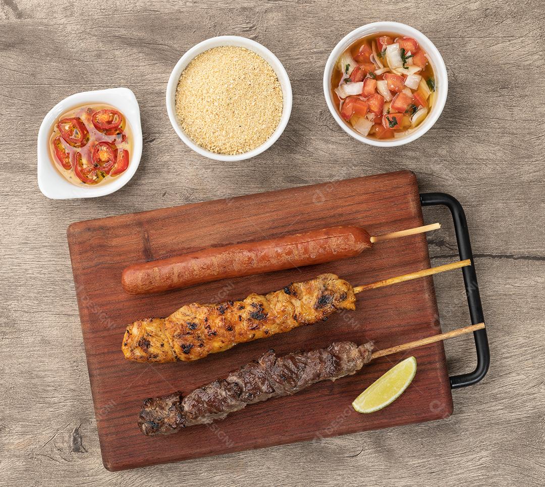 Espetos de carne, frango e salsicha de porco sobre tábua de madeira com farofa e vinagrete.
