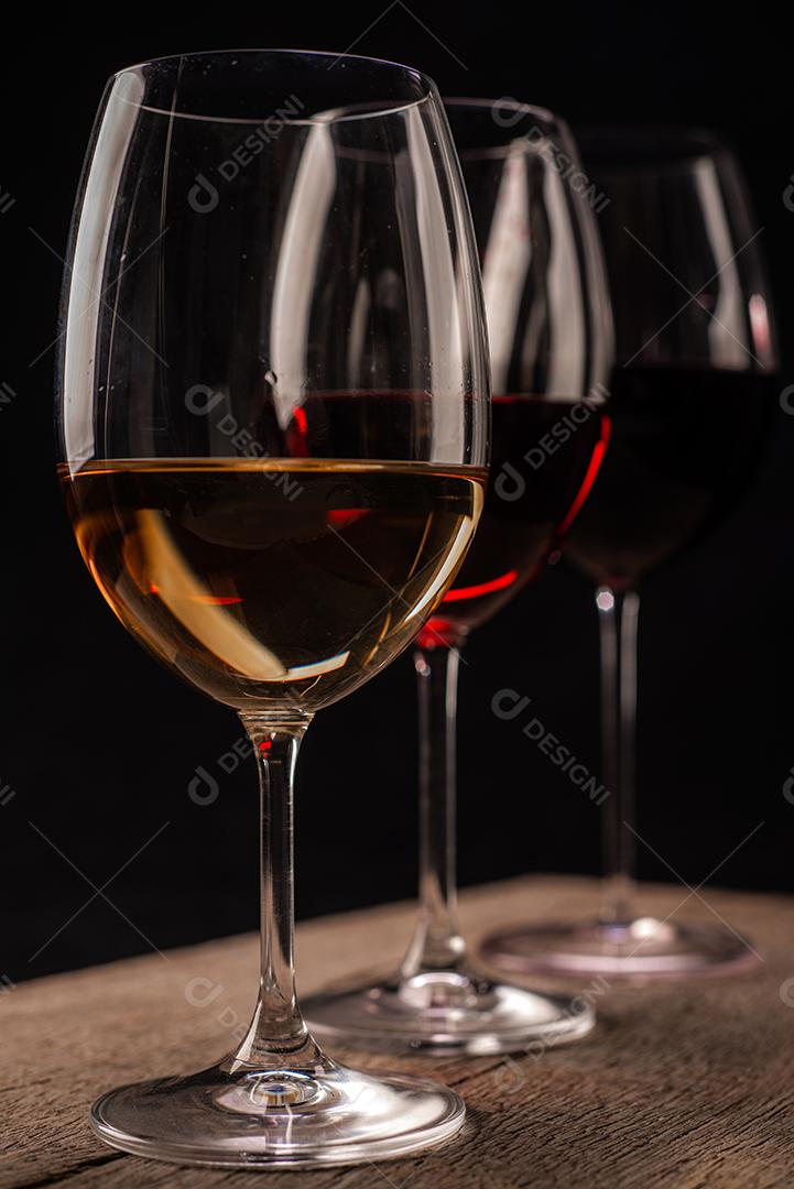 Taças de cristal com vinho tinto e branco sobre a mesa de madeira rústica em fundo preto.