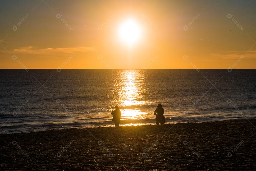 Pôr do sol na praia com sol no céu e 2 pessoas em silhueta.
