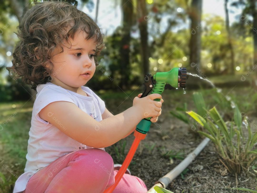 Menina brasileira bonitinha se divertindo brincando com mangueira de água no jardim.