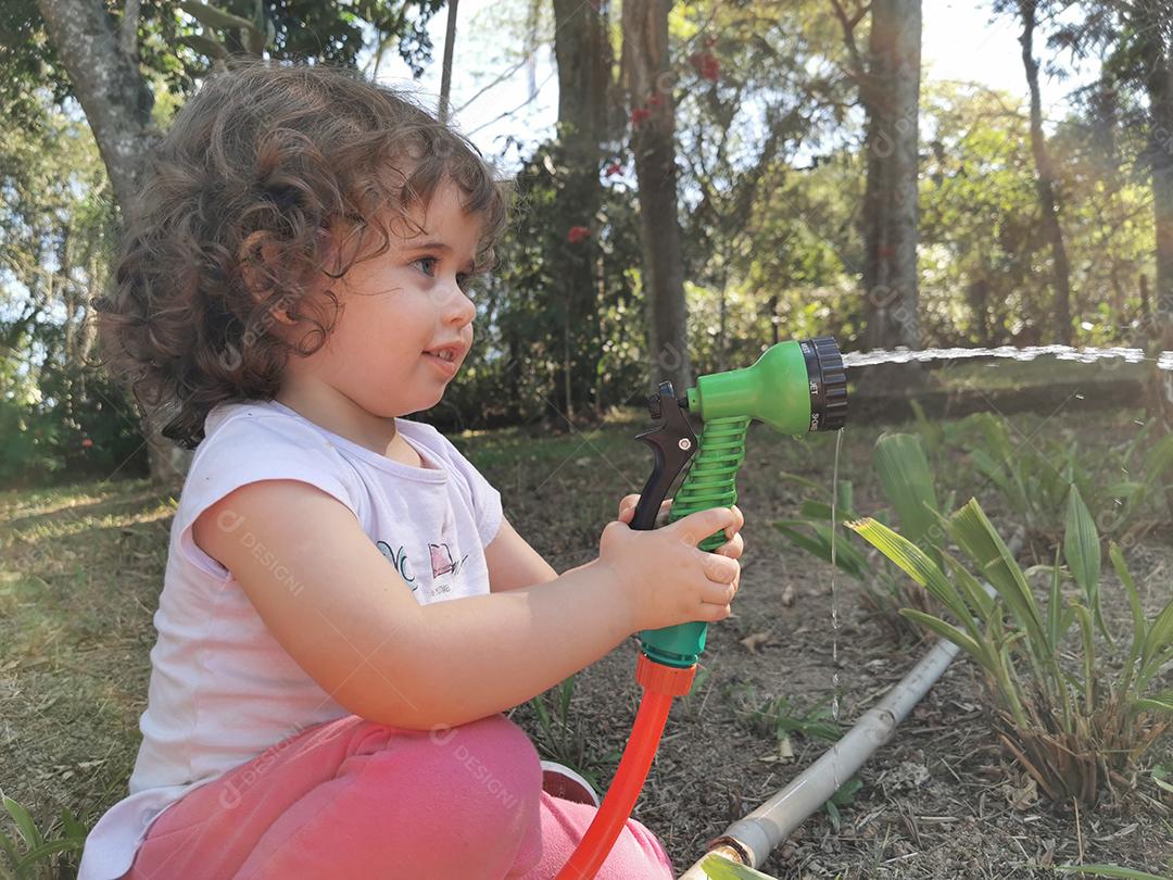 Menina brasileira bonitinha se divertindo brincando com mangueira de água no jardim.