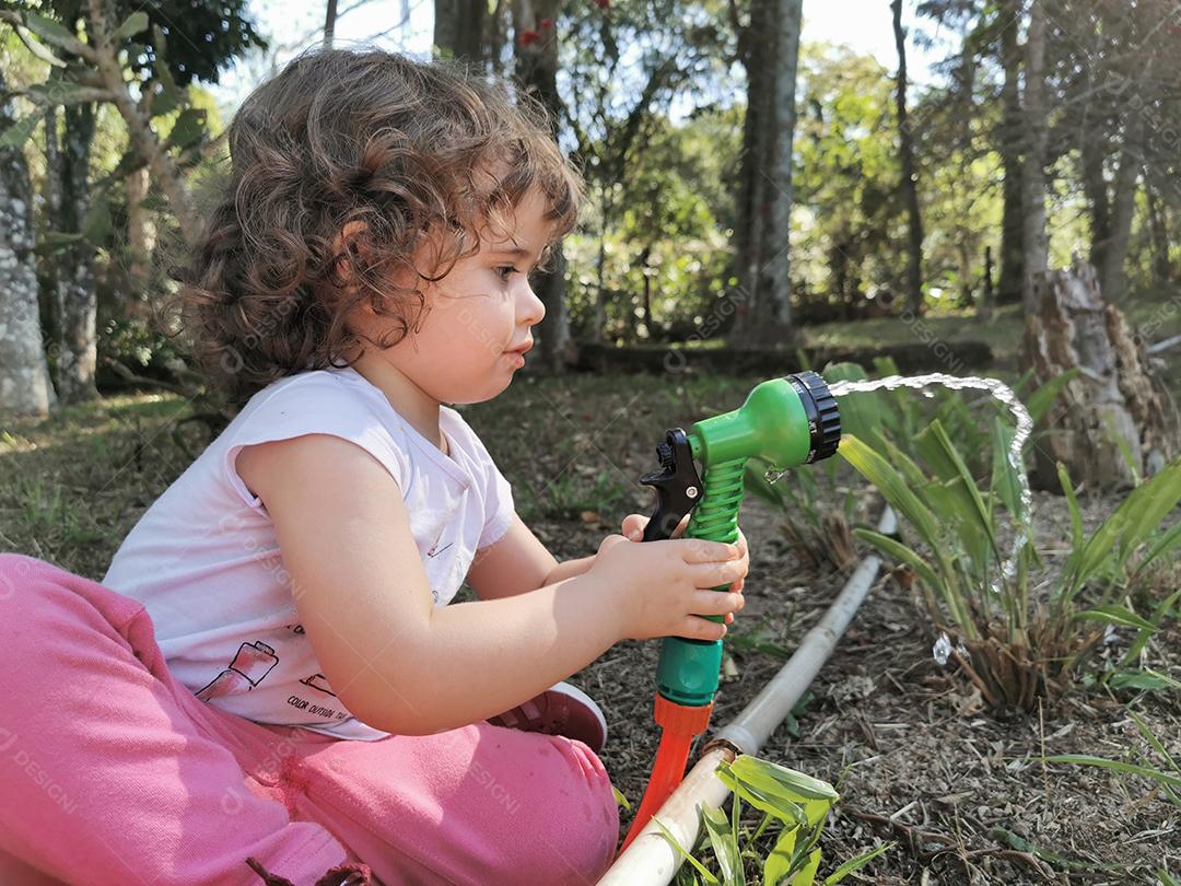 Menina brasileira bonitinha se divertindo brincando com mangueira de água no jardim.