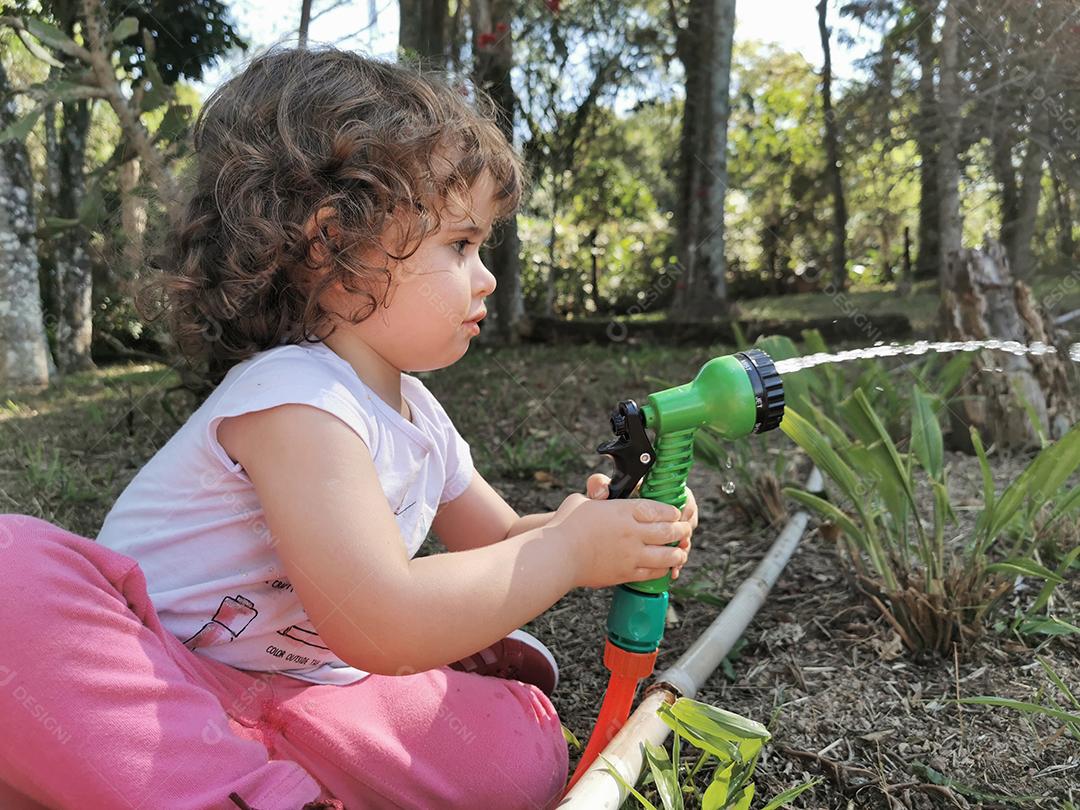 Menina brasileira bonitinha se divertindo brincando com mangueira de água no jardim.