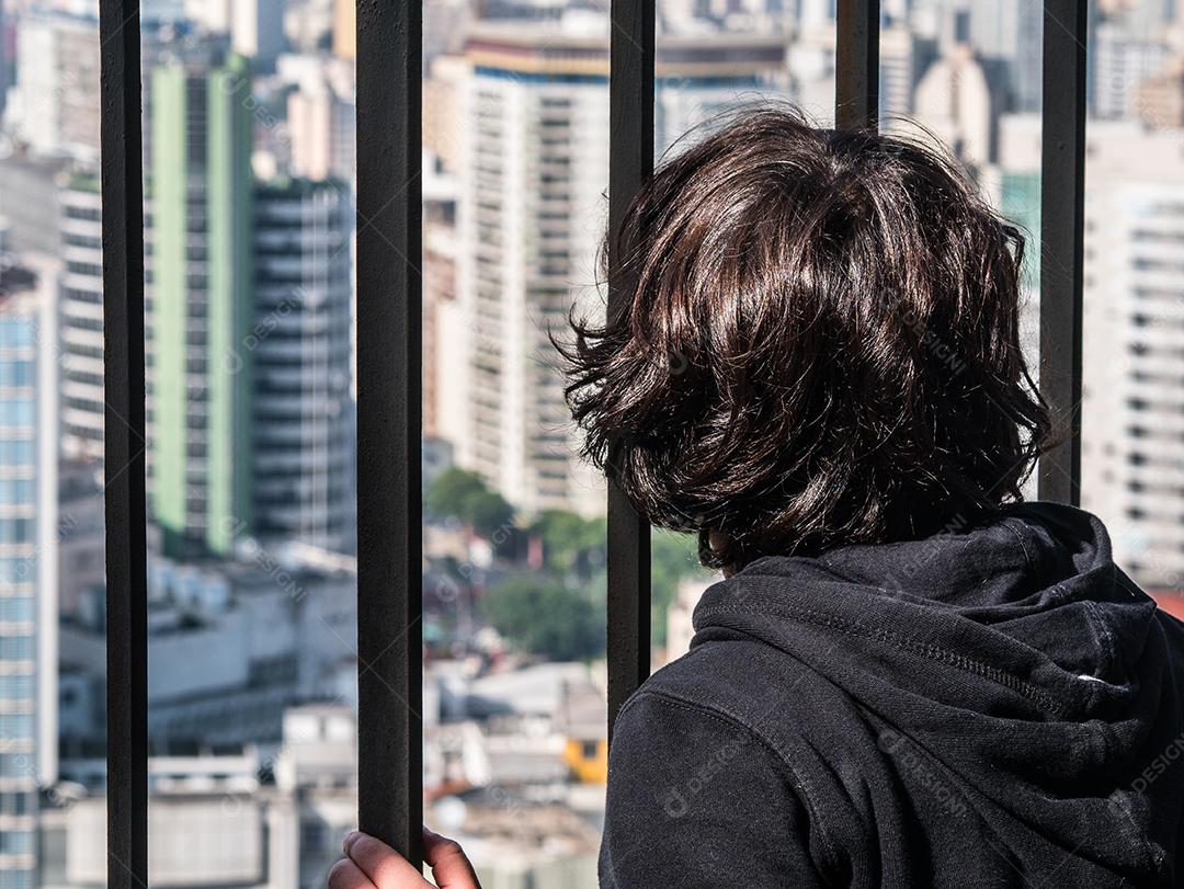 Menino olhando para a cidade atrás das grades.