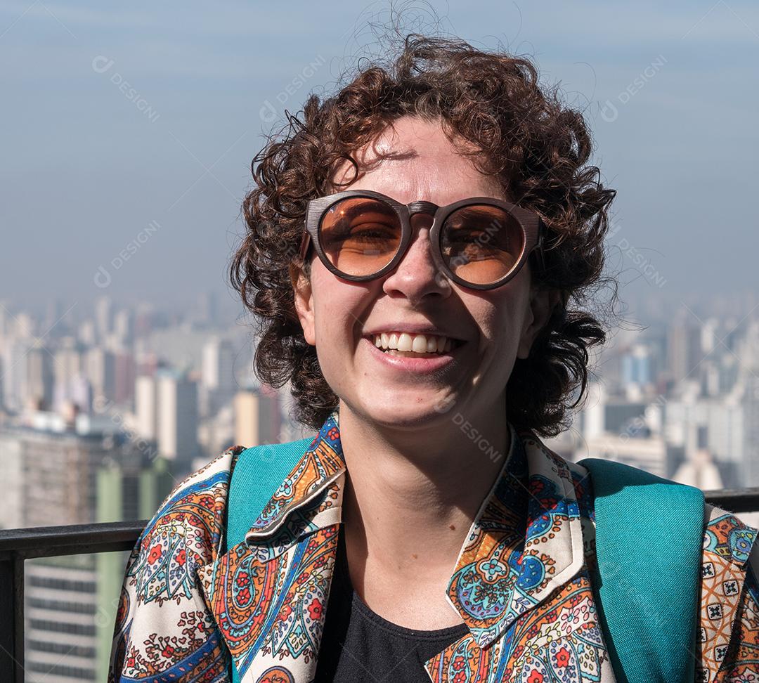 Mulher bonita e moderna posando para a câmera usando óculos escuros com a cidade de São Paulo em segundo plano.