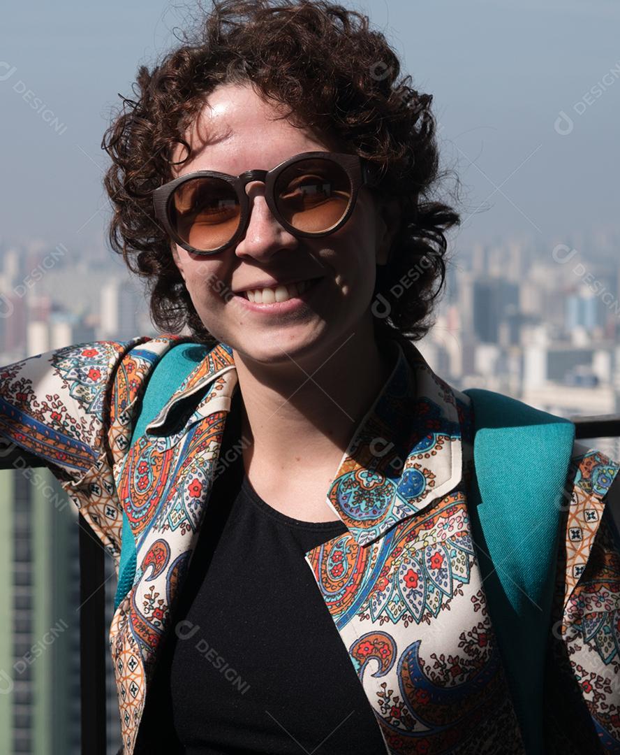 Mulher bonita e moderna posando para a câmera usando óculos escuros com a cidade de São Paulo em segundo plano.