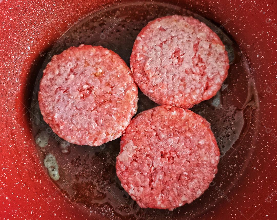 Três hambúrgueres sendo fritos em uma frigideira vermelha..