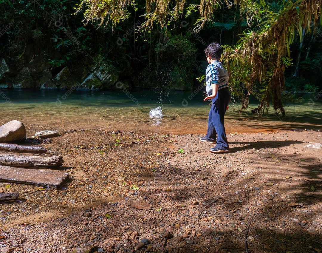 Menino jogando pedra no rio na floresta tropical.