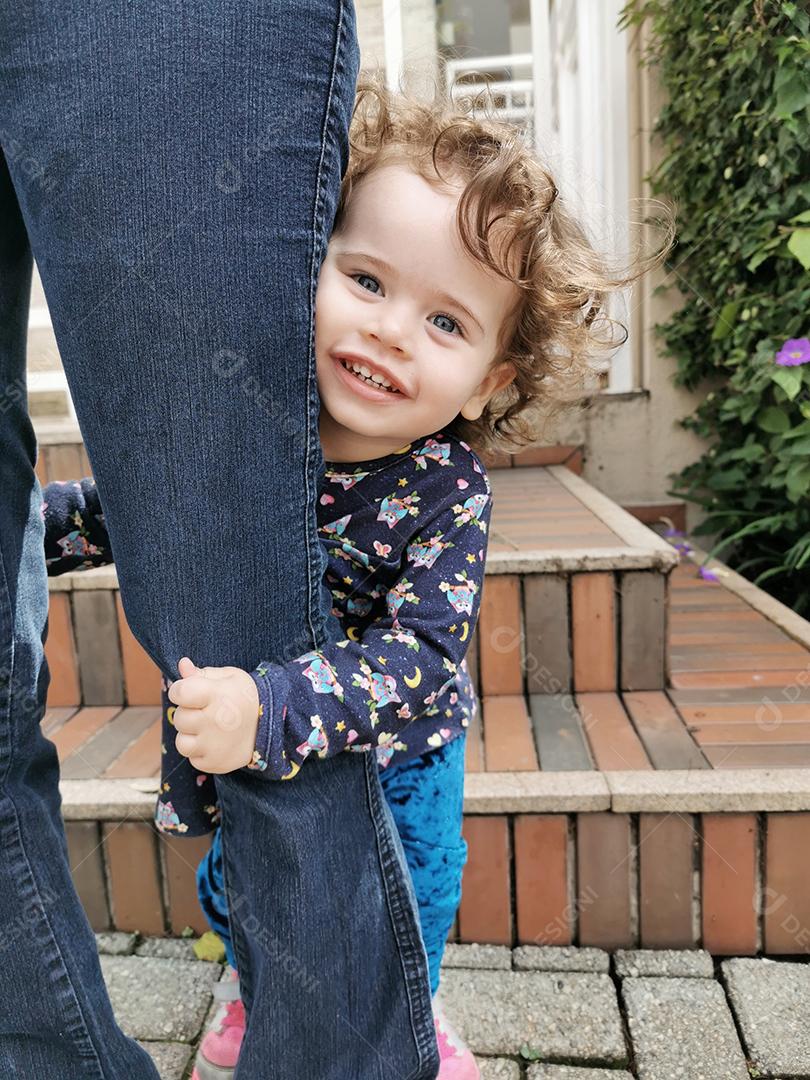 Linda garotinha segurando a perna da mãe e sorrindo.