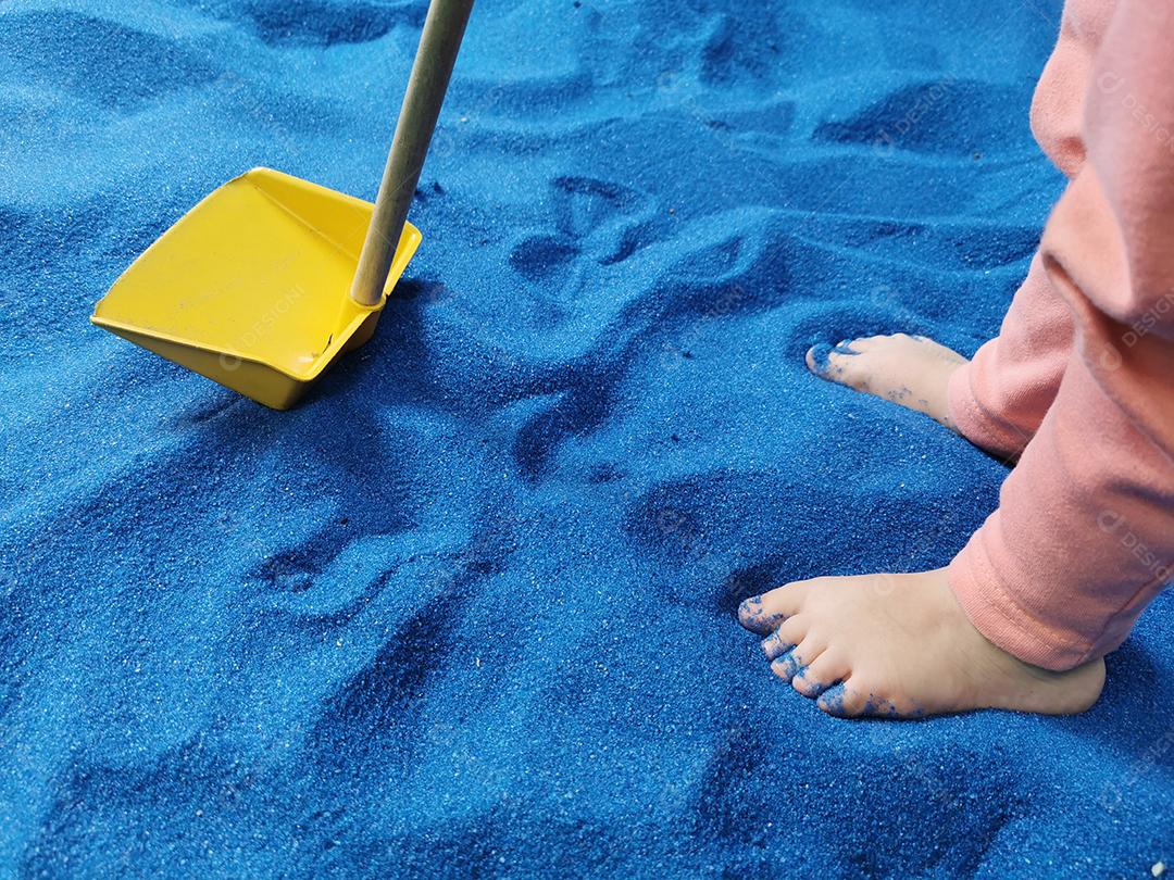 Pés de criança pequena brincando com pá na areia azul.