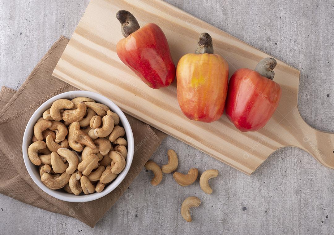 Três cajus sobre uma mesa de madeira ao lado de uma tigela de castanhas de caju brasileiras.
