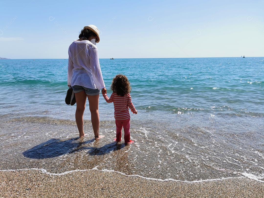 Mãe e filha de mãos dadas com os pés descalços em frente à praia