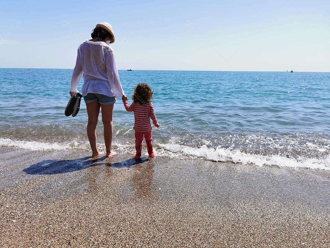 Mãe e filha de mãos dadas com os pés descalços em frente à praia