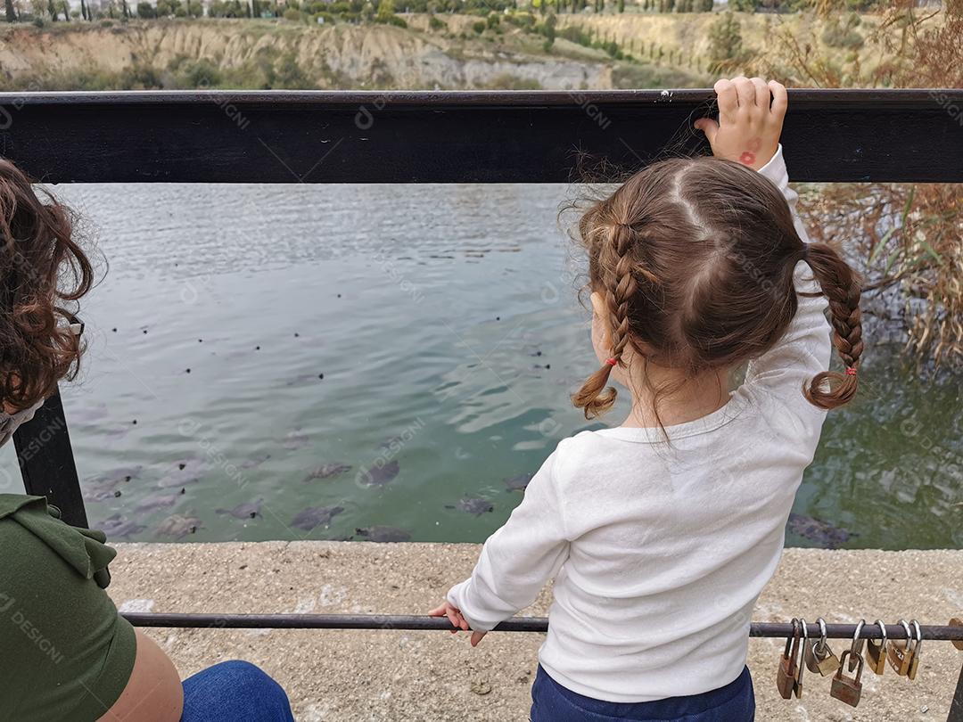 Mãe e filha de costas olhando tartarugas no lago.