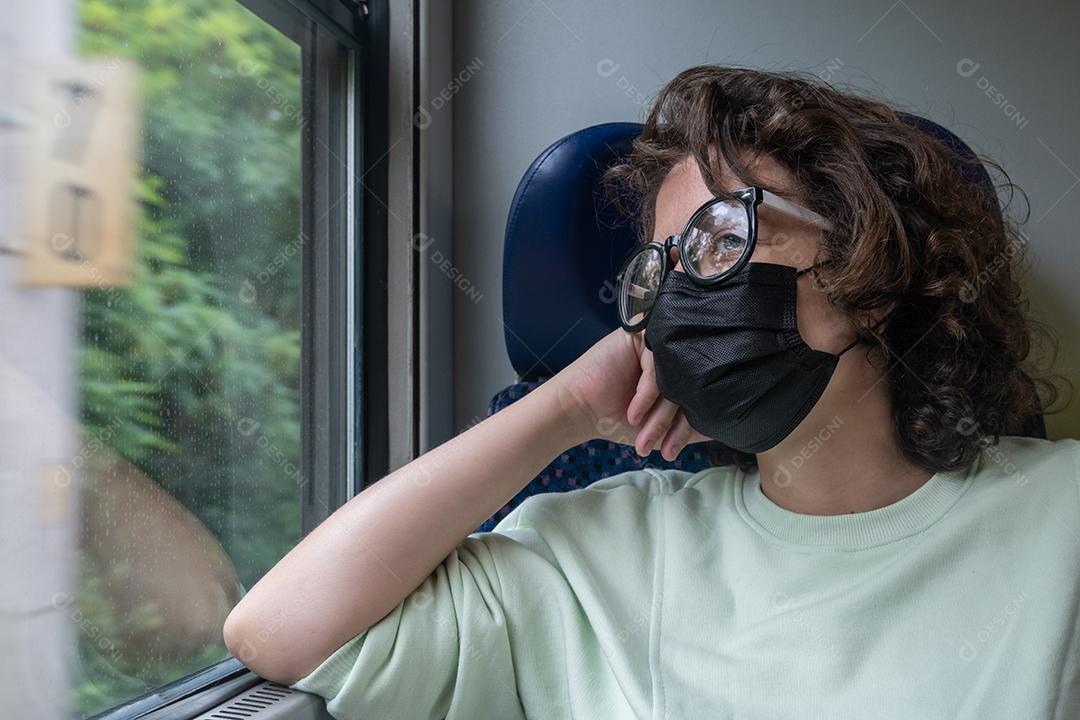Vista de uma jovem viajando de trem durante a pandemia usando óculos e máscara preta.